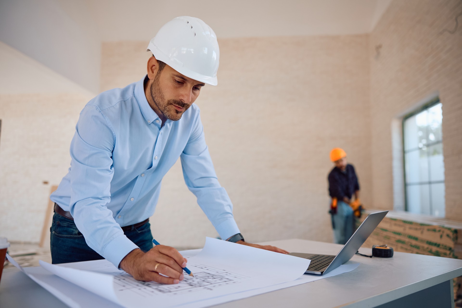 An engineer in a white hard hat reviews blueprints at a construction site.