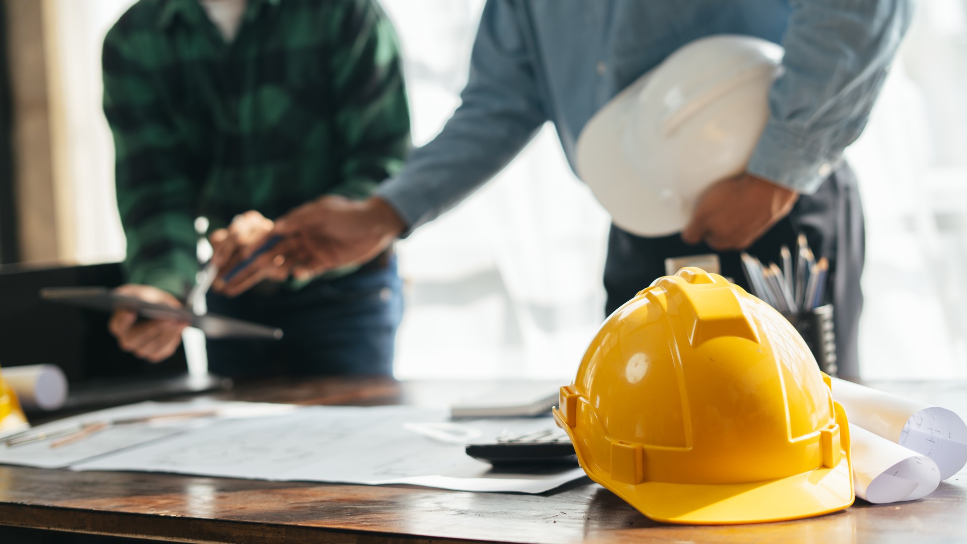 A yellow hard hat on a table with blueprints, as two workers discuss construction plans.